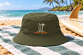 Beach Bound Bucket Hat shown lying on a striped beach towel next to a surfboard on golden sand
