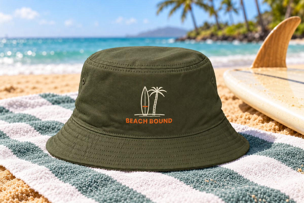 Beach Bound Bucket Hat shown lying on a striped beach towel next to a surfboard on golden sand
