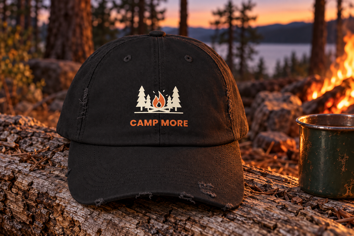 Camp More Distressed Cap shown sitting on a weathered wooden log next to a small glowing campfire in a pine forest at dusk