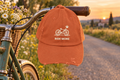 Ride More Distressed Cap shown hanging on the handlebar of a vintage bicycle parked on a quiet countryside road lined with wildflowers