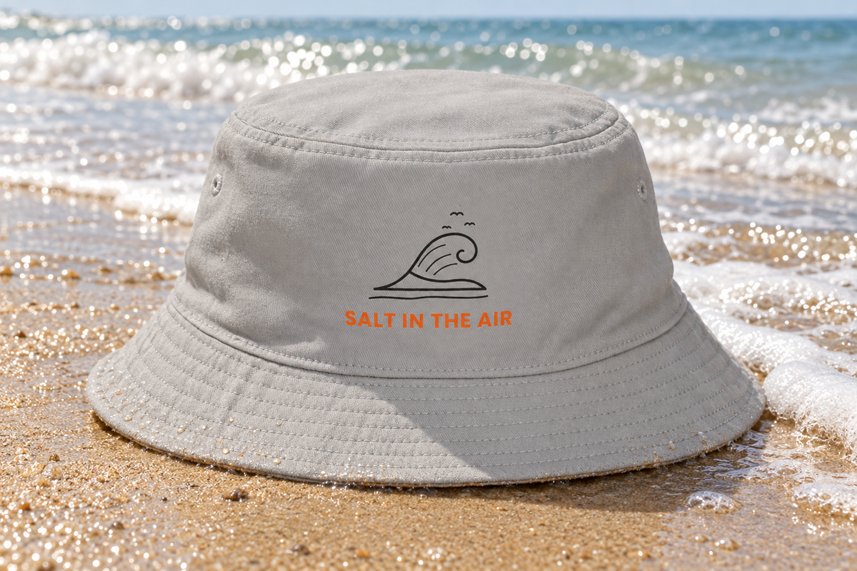 Salt In The Air Bucket Hat shown lying on smooth wet beach sand near the shoreline with gentle waves and sea foam
